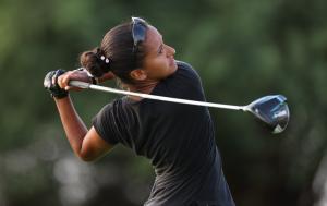 A young, tan complected female golfer with brown hair in a back bun, wearing all black and swinging a golf club.