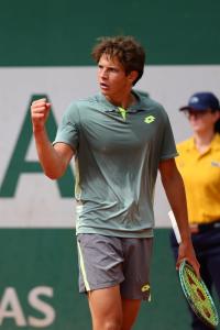 A tall male with brown hair in a green tennis shirt and grey shorts, holding a racket in his left hand while pumping his right fist.