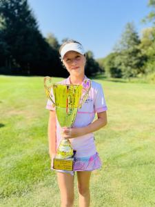 A young woman on a green field, holding a huge gold trophy. She's smiling at the camera, with her blonde hair pulled back and a white visor and wearing a pink short sleeved golf shirt with a multicolor skirt.