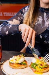 A person in a floral top cuts into an Eggs Benedict with avocado and smoked salmon, topped with a runny yolk, pomegranate seeds, and sprouts, served on a white plate at Ayza Wine & Chocolate Bar.