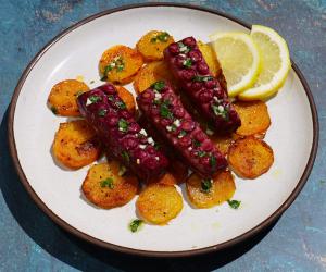 A white plate with three slices of plant-based octopus by Revo Foods, garnished with chopped garlic and herbs, served over golden roasted potato slices and lemon wedges on a textured blue background.