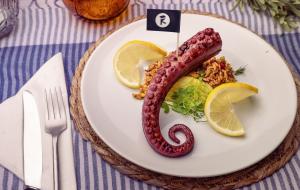 A plated dish featuring a plant-based octopus tentacle by Revo Foods, served with lemon wedges, microgreens, and a bed of spiced grains, styled on a white plate with a striped blue tablecloth.