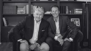 The photo is black and white , Martin Kelly on left with sandy grey hair wears a light blue and smiles at the camera, Justin Bulley is seated to his right in a light grey suit