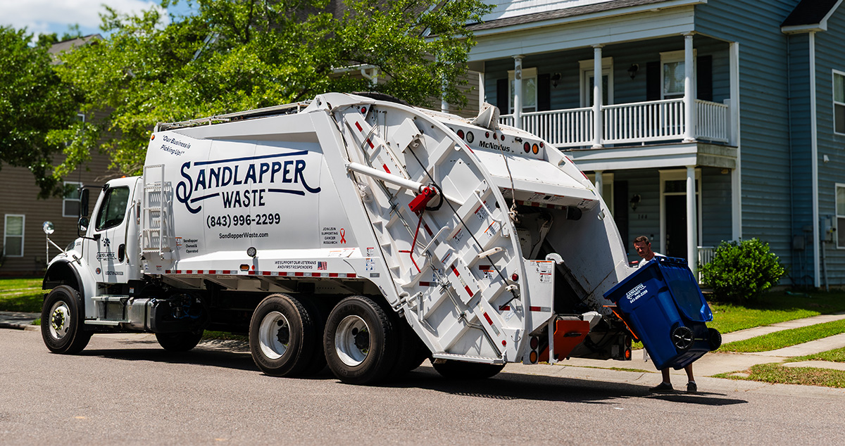 Residential garbage collection by Sandlapper Waste truck in suburban neighborhood, with worker handling blue recycling bin near curbside in front of modern two-story house.