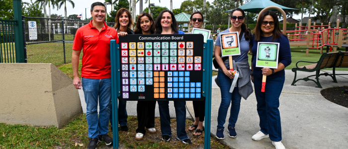 A group of people stands beside a colorful communication board in a park in Village of Wellington, holding signs with pictograms for basic needs.