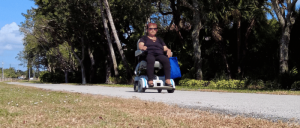 A person riding a mobility scooter along a paved path lined with trees, carrying a blue bag, enjoying a sunny day.