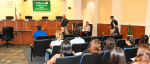 A speaker addresses an audience in a conference room, with panelists seated in front and a presentation displayed in the background.