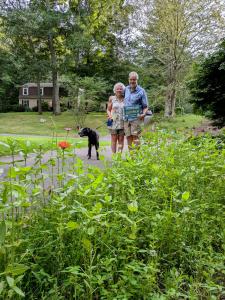 A Connecticut couple and their dog, Bella, stand in front of a pollinator-friendly strip they planted for Reduce Your Lawn Day, holding a sign. Their mini meadow attracts butterflies and hummingbirds, showing how small spaces can support biodiversity.