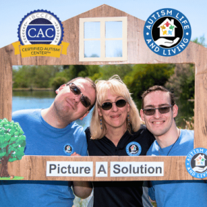 Three smiling individuals hold a wooden frame shaped like a house, promoting the "Picture A Solution" initiative near a tranquil lake.