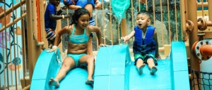 A woman and a young boy slide down vibrant blue water slides, splashing water in a fun indoor water park setting.