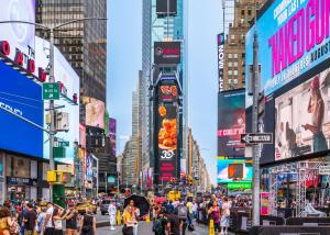 A large digital billboard in Times Square features bb.q Chicken’s 30th anniversary ad with crispy Korean fried chicken, as crowds gather below.