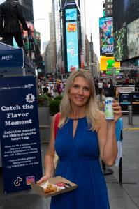A woman holds a can of Milkis and a tray of bb.q Chicken at the Times Square event, with campaign signage and digital billboards visible in the background.
