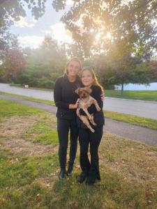 The founder of DogSol, Jackie Jolie stands next to Kara who is holding Marley. Kara is the owner of the rescued Yorkie, Marley, who is medium-sized and has brown and black fur. The three of them are standing outside with the sun shining behind them in a y