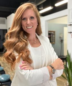 Dr. Brittany Tinker stands proudly, arms crossed, wearing white while her office is slightly visible in the background. She smiles warmly.