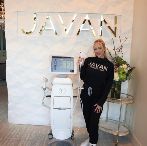 A smiling woman stands in front of a textured white wall with the gold-lettered "JAVAN" logo, wearing a black “JAVAN FOREVER YOUNG” sweatshirt. She is holding a handheld applicator attached to a modern aesthetic treatment machine with a touchscreen interf