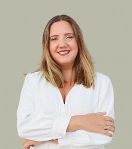 Smiling woman with straight shoulder-length light brown hair, wearing a white blouse, standing confidently with arms crossed against a plain, muted green background.