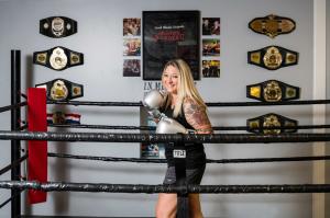 Professional Kickboxer and Fitness Trainer Christina Rondeau standing in a boxing ring with boxing gloves on and her Champion Belts are on a wall behind the ring