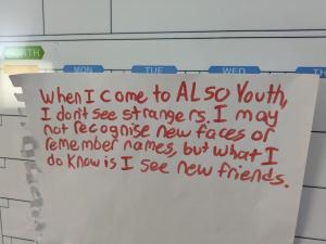Handwritten sign in red marker that says: "When I come to ALSO Youth, I don’t see strangers. I may not recognise new faces or remember names, but what I do know is I see new friends." The sign is taped to a whiteboard calendar.