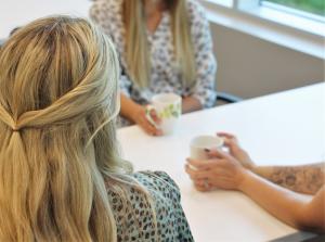 Three individuals, two of them holding coffee cups, sit around a table together. Their faces are not visible, but they appear to be engaged in conversation.