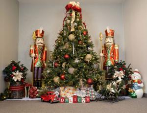 "Elegant indoor Christmas display with a decorated tree in red, gold, and silver ornaments, flanked by two large nutcracker statues, surrounded by wrapped gifts, festive floral arrangements, a red toy truck, and a snowman figurine."