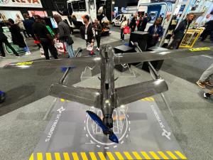 Overhead view of a carbon fibre fixed-wing VTOL drone on display at a trade exhibition, surrounded by attendees and security vehicles.