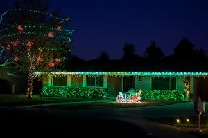 Scottsdale home with green-lit hedges, red light spheres in the tree, icicle roofline lights, and an illuminated sleigh filled with gifts. Designed by Margie Mae’s Holiday Decor to deliver nostalgic holiday beauty with a modern desert glow.