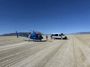 A blue Care Flight helicopter and a Royal Ambulance vehicle meet in the Black Rock Desert for a patient transfer, with EMS crews walking across cracked dry earth under a clear blue sky.