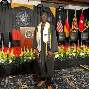 Michael Ameer wearing a graduation gown and cap at the University of Maryland Global Campus Europe ceremony, standing on a stage decorated with flags and flowers