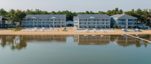 Beachfront hotel with multiple levels, lounge chairs on sandy shore, and calm water reflecting the buildings and surrounding trees.
