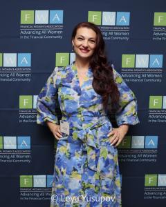 a woman with long dark hair wearing a blue and green dress stands in front of a navy backdrop that is dotted with the FWA logo
