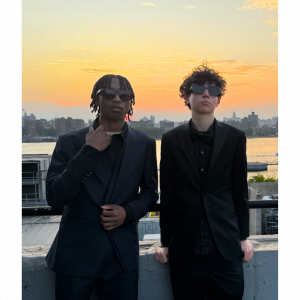 Two young men in black suits and sunglasses, standing in front of the sunset with the New York City skyline behind them. Beckett Zahedi on the left, Derrick Webster on the right—co-founders of Realer Estate.