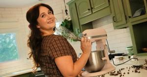 Missy Lanning smiling confidently while holding a stainless steel whisk in front of her pink mixing bowl. She is standing in her Tennessee farmhouse kitchen with green cupboards and green plants hanging in the light of the windows.