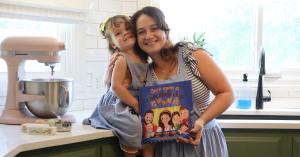 Missy Lanning smiles while her young daughter Brielle hugs her in a cozy kitchen. Both are wearing matching blue aprons with Once Upon A Cookie logos on them.
