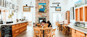 Bright, modern cafe interior featuring wooden tables, a stone fireplace, artwork on the walls, and patrons enjoying their time.
