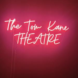 A glowing pink neon sign that reads “The Tom Kane THEATRE” against a dark background. The sign is written in a combination of cursive and capital letters, creating a vibrant and theatrical visual effect.