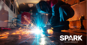 A welder in a protective mask kneels while working on a metal floor, creating a bright shower of blue and orange sparks.