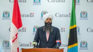 The honorable Randeep Sarai, Secretary of State for International Development, Government of Canada, speaking in Tanzania. He is standing behind a podium wearing a dark suit, light blue shirt and pink tie. The Canadian flag is on the left of the podium an