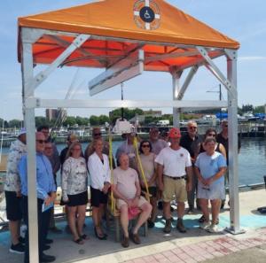 Group of community members, city officials, and Handicaptain team posing around the newly installed ADA-compliant Handicaptain Boarding Station at Dunkirk Pier, with a man seated in the lift demonstrating its use.