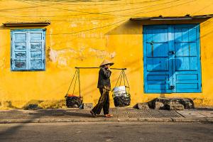 Vietnamese street vendor carrying baskets with a shoulder pole in front of a vibrant yellow wall and blue windows in Hoi An, captured by Réhahn.