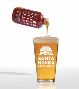 A bottle of Bees Knees Spicy Honey is being poured into a pint glass of beer labeled "Santa Monica Brew Works" on a white background.
