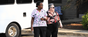 Two women walk together outside, one in a floral scrub top and the other in a patterned shirt, with a white bus in the background.