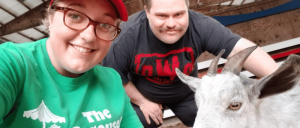 Two people smiling in a barn setting, leaning close to a curious goat, showcasing a friendly interaction with the animal.