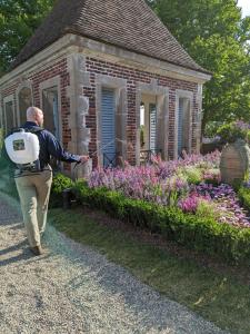 The image shows a Deer Solution technician applying deer repellent to a beautifully landscaped garden. The technician is wearing khaki pants, a dark long-sleeve shirt, and boots, with a white Deer Solution-branded backpack sprayer on his back. He is spray