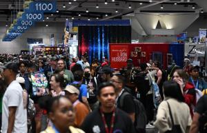 A long shot showing a packed hall, the aisle numbers, and in the distance, the bright red Galaxy Press booth shining bright at San Diego Comic Con 2025.