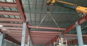 Workers installing steel beams using a crane inside a pre-engineered building under construction at an industrial site.