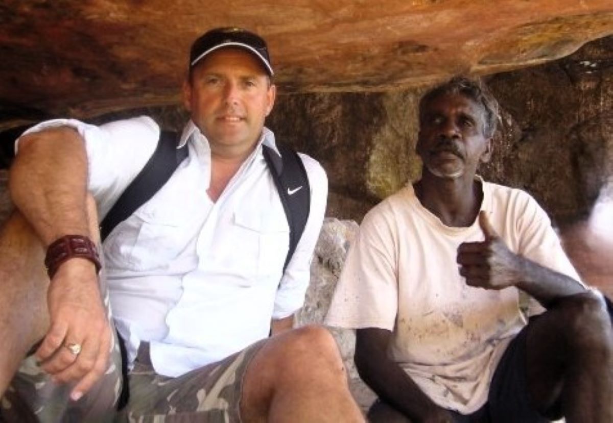 Paul Rushworth-Brown sitting beside an Aboriginal Elder giving a thumbs-up under a rock shelter in Arnhem Land.