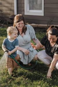 Two mothers smiling while playing with their young son under a garden hose on a warm day