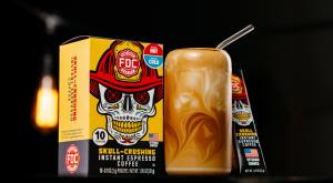 Box of Fire Department Coffee Skull-Crushing Instant Espresso Coffee beside a glass of swirled coffee with a straw. The design features a skull with a firefighter helmet.