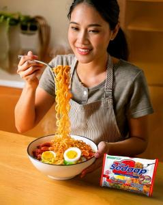 Smiling woman lifting noodles from a bowl of Mi Sedaap Original Fried Noodles, garnished with boiled egg and vegetables, with the product pack displayed in the corner.