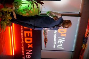 Dr. Mohana Rao Patibandla speaking on stage at the TEDx Nerul event, wearing a blue vest and white shirt, holding a microphone, with a red neon sign and green plants in the background. The backdrop features the TEDx Nerul logo and the text "X=independentl